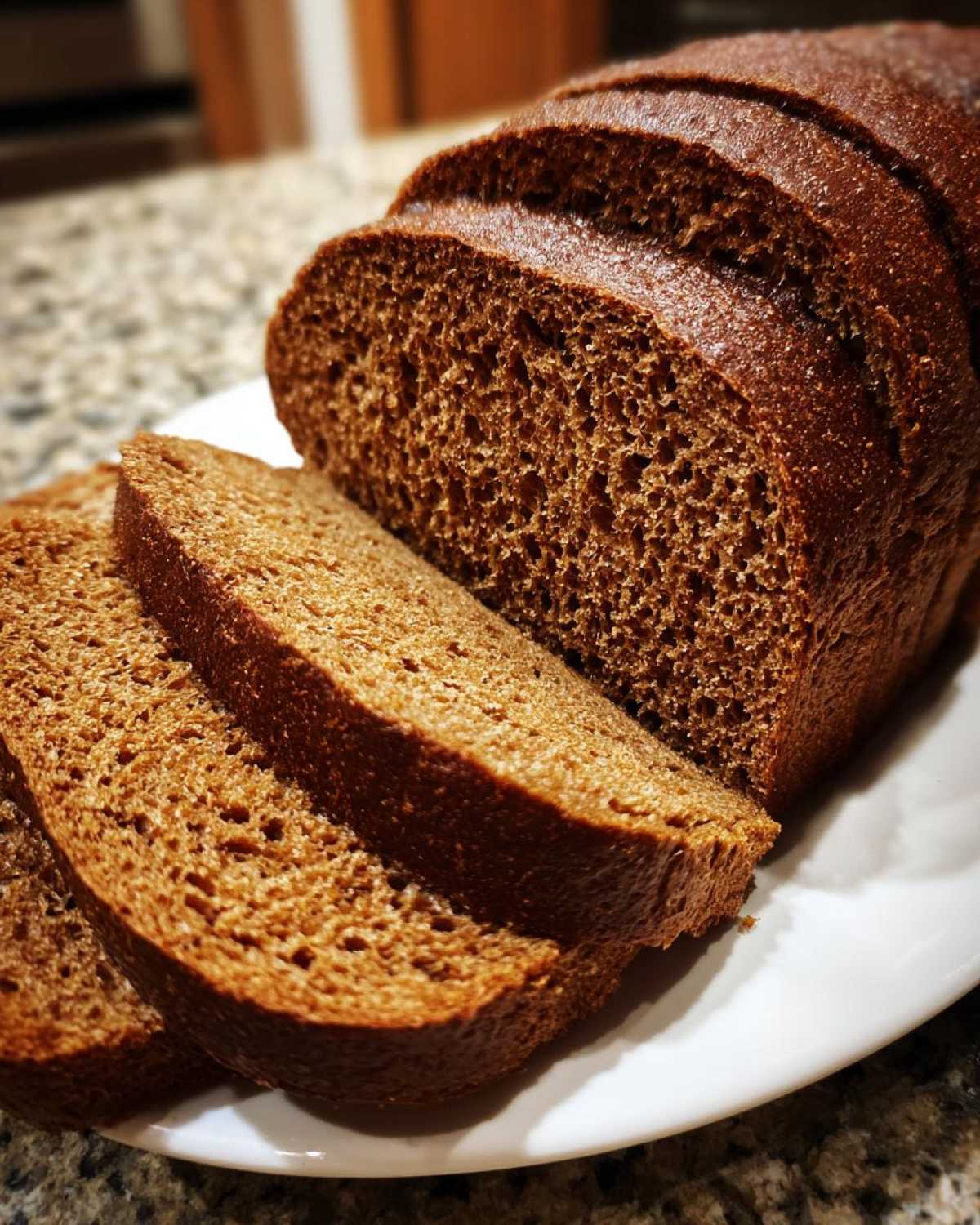 Close-up of a sliced loaf of rich, dark brown honey wheat bread on a white plate.