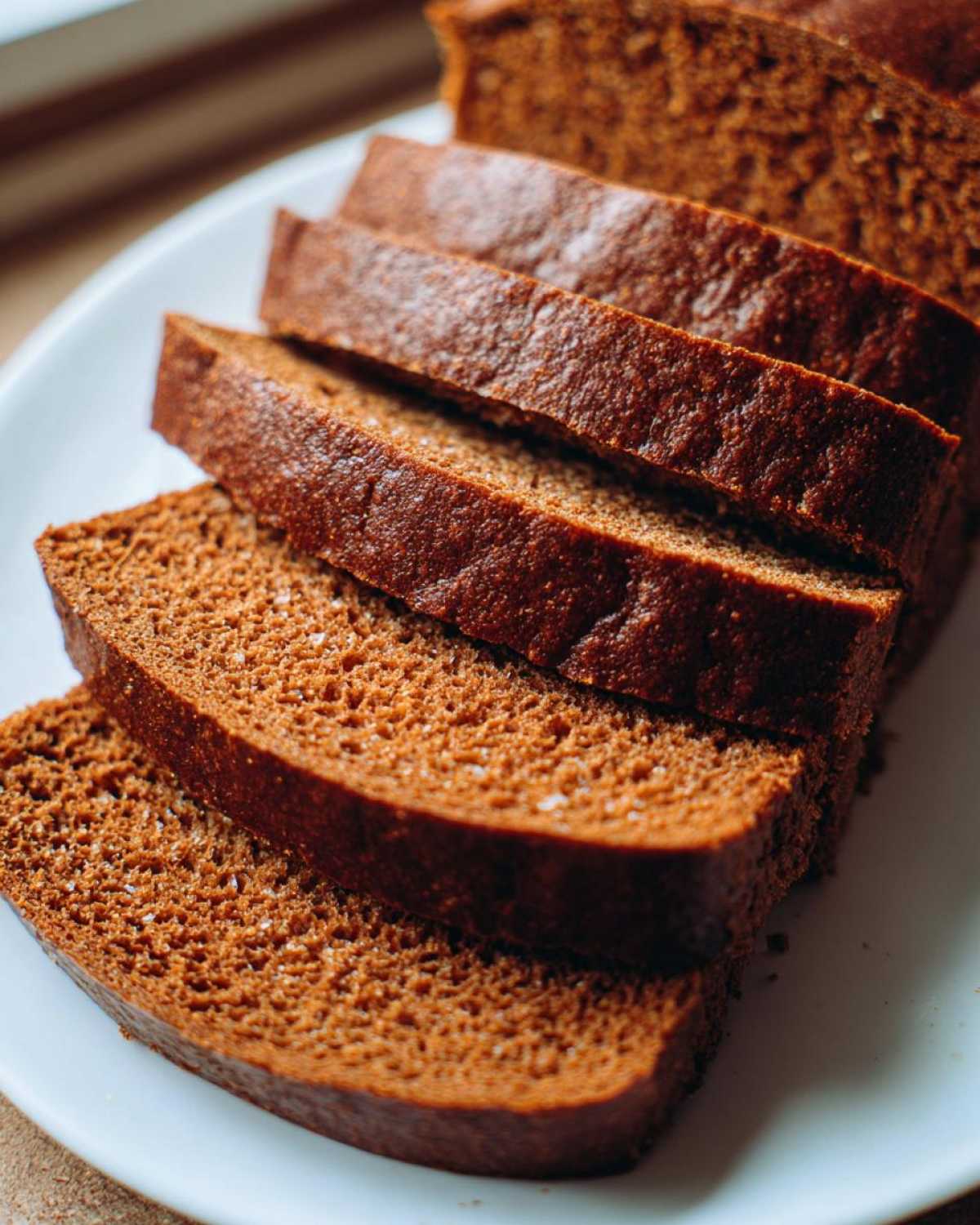 Close-up of several thick slices of rich, dark brown honey wheat bread stacked on a white plate.