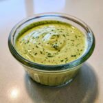Close-up of thick, pale green Copycat Green Goddess Ranch Dressing topped with finely chopped herbs in a small glass ramekin.