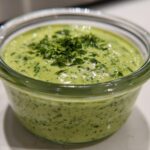Close-up of creamy, bright green Copycat Green Goddess Ranch Dressing topped with fresh chopped herbs in a small glass container.