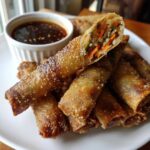 A pile of crispy, golden brown Sesame Veggie Spring Rolls served with a small white bowl of dark dipping sauce.