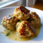 Four golden-brown Easy Creamy Greek Chicken Meatballs resting in a pool of light, creamy sauce, garnished with fresh parsley.