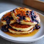 A close-up of a stack of three fluffy Blueberry Pancakes topped with cooked blueberries and drizzled with syrup.