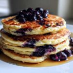 A tall stack of three fluffy Blueberry Pancakes topped with fresh blueberries and drizzled with syrup.