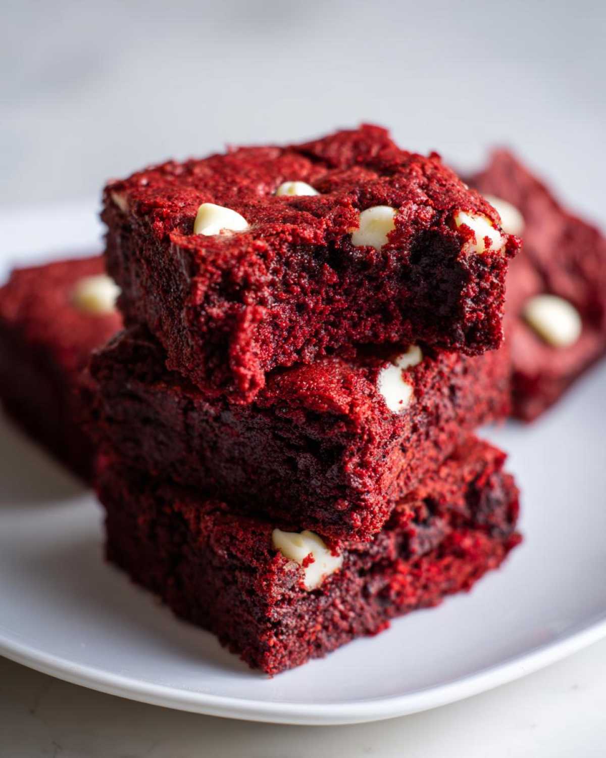 A stack of three fudgy red velvet brownies studded with white chocolate chips, one on top has a bite taken out.