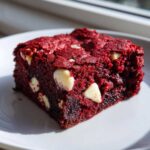 A single, rich, fudgy red velvet brownie square studded with white chocolate chips on a white plate.