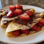 Two folded Gluten-Free Chocolate Strawberry Crepes topped with melted chocolate and fresh sliced strawberries.