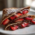 Stack of folded Gluten-Free Chocolate Strawberry Crepes oozing with chocolate sauce and fresh strawberries, dusted with powdered sugar.
