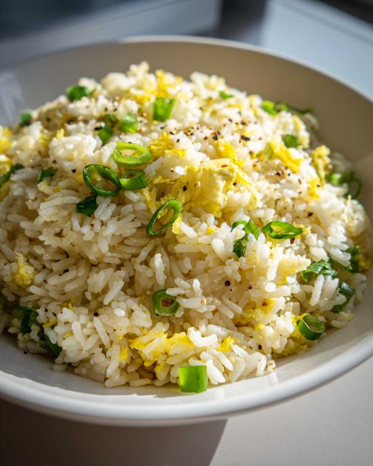 Close-up of a white bowl filled with Gluten-Free Lemon Pepper Fried Rice, featuring scrambled egg pieces and sliced green onions.