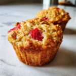 A close-up of a freshly baked Gluten-Free Strawberry Oat Muffin showing oats and a bright red strawberry piece on top.