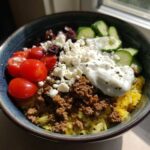 A vibrant Greek Ground Beef Rice Bowl featuring seasoned meat over yellow rice, topped with tzatziki, feta, tomatoes, cucumbers, and olives.