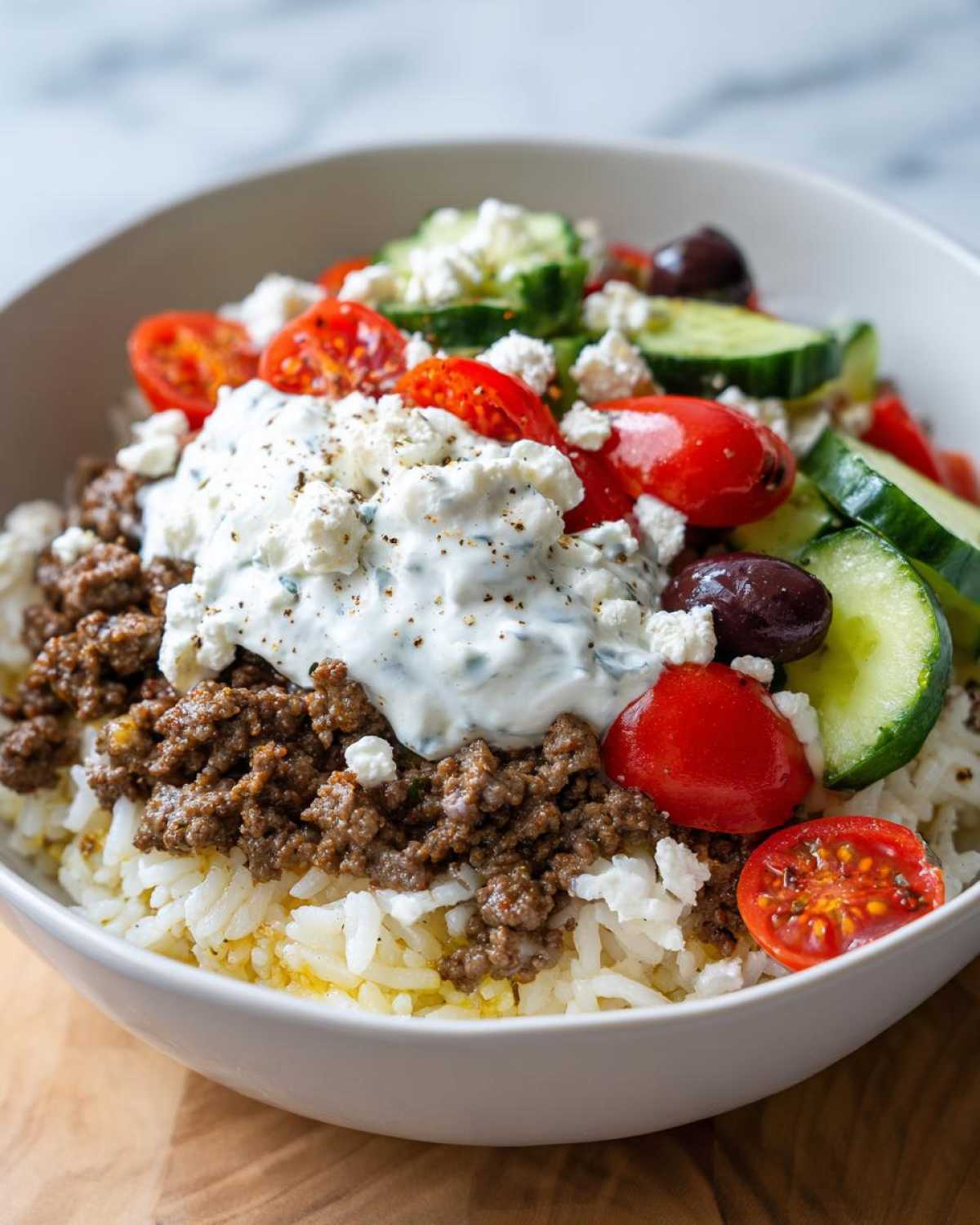 Close-up of a Greek Ground Beef Rice Bowl topped with tzatziki, feta, tomatoes, cucumbers, and olives.