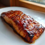 A beautifully caramelized fillet of Honey Soy Glazed Salmon resting on a white plate with excess glaze.