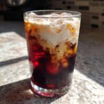 Close-up of a tall glass filled with Iced Cherry Vanilla Cold Brew, ice, and a layer of cream swirling into the dark coffee.