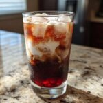 A tall glass of Iced Cherry Vanilla Cold Brew with ice cubes and cream swirling into the dark coffee.