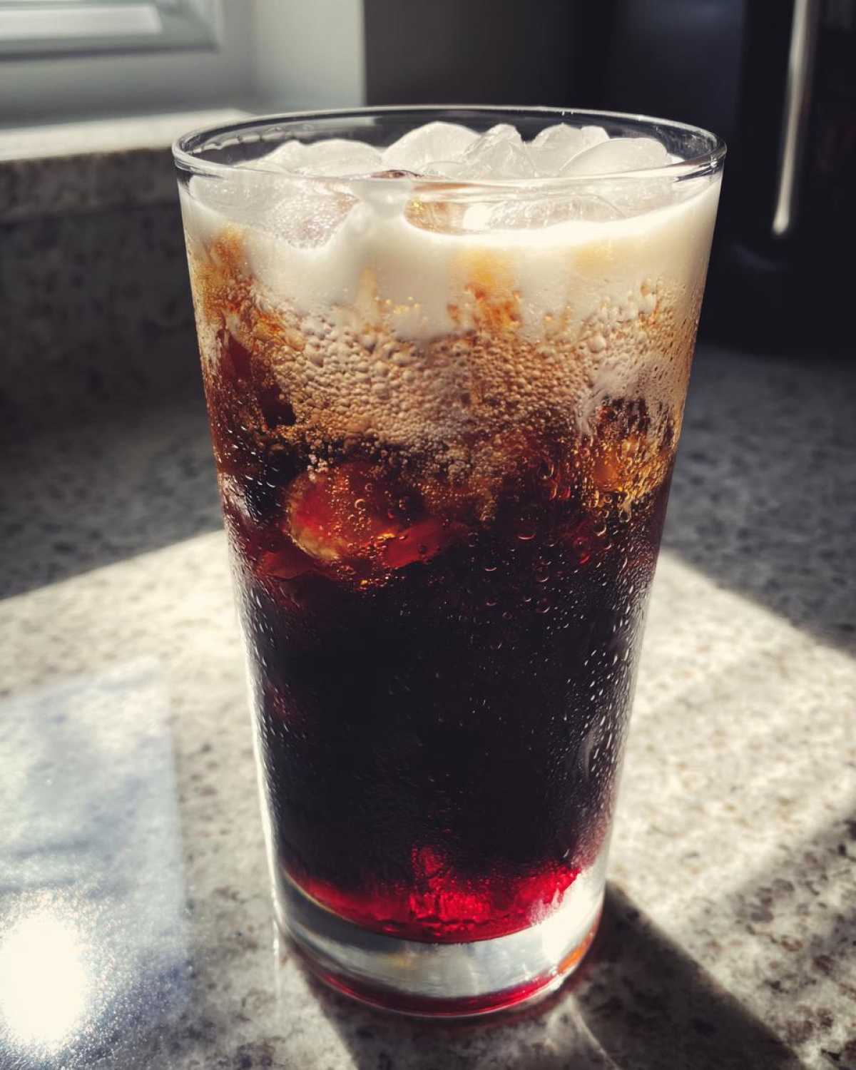 A tall glass of Iced Cherry Vanilla Cold Brew with ice and a creamy foam top, sitting on a sunlit counter.