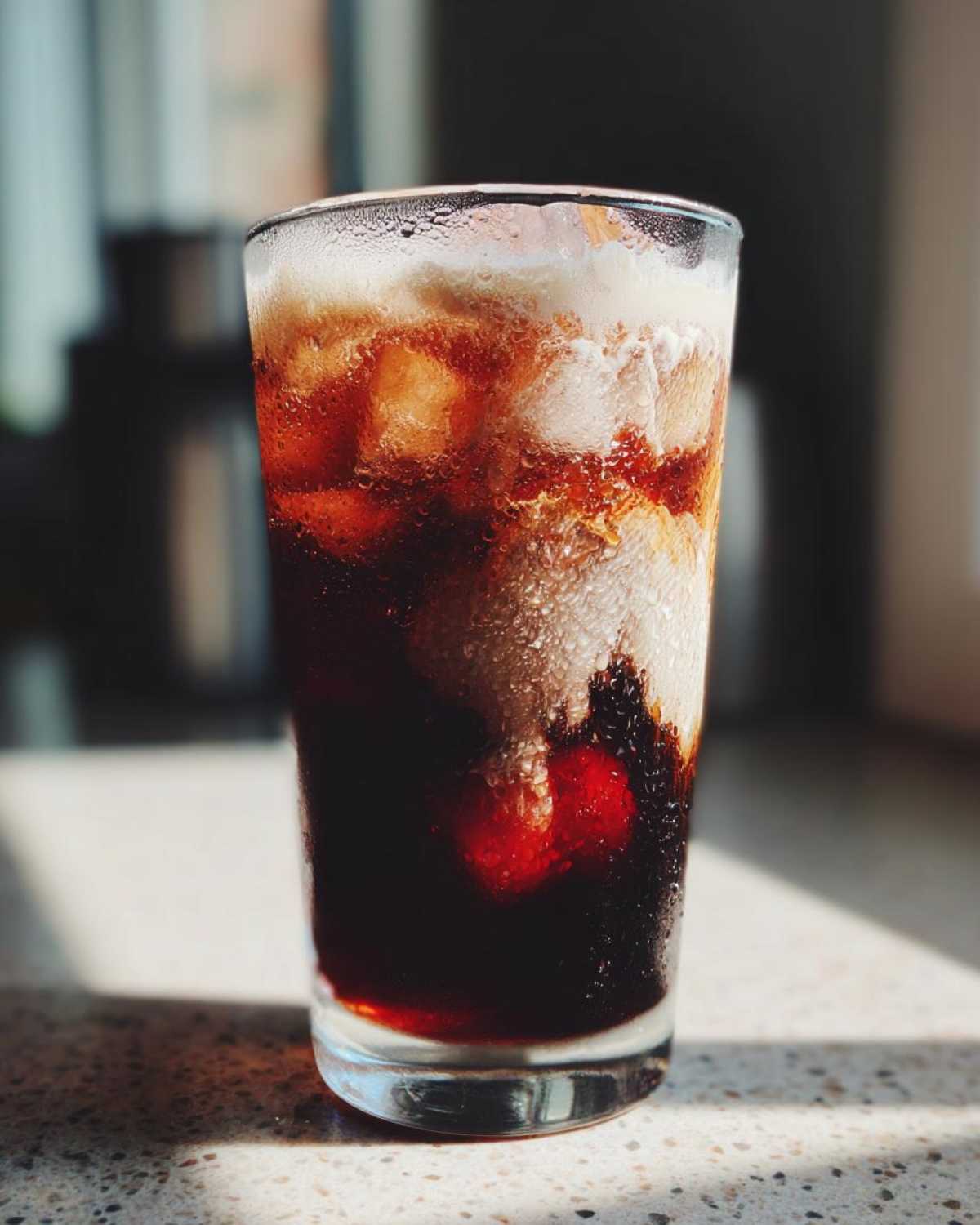 A tall glass filled with Iced Cherry Vanilla Cold Brew, showing dark coffee, ice, and a creamy layer on top, glistening with condensation.