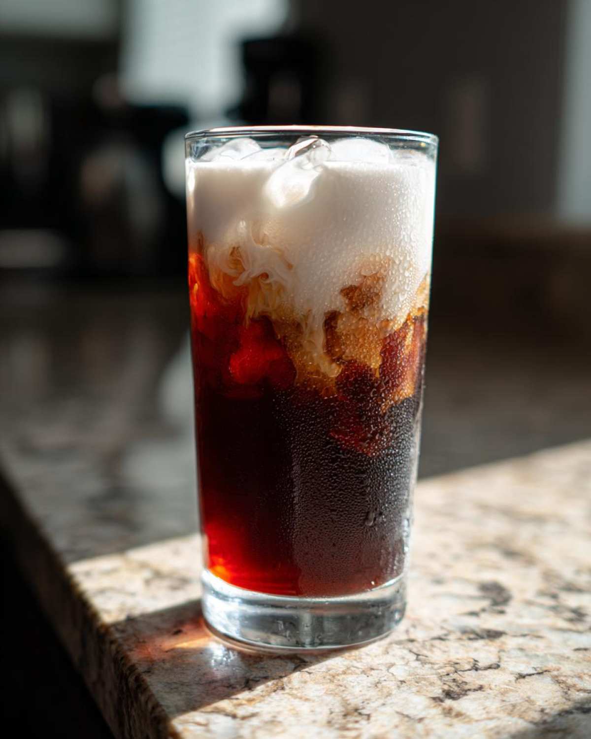 A tall glass of Iced Cherry Vanilla Cold Brew showing dark coffee, red cherry syrup, and white cream swirling on top over ice.