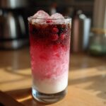 A tall, condensation-covered glass filled with Iced Raspberry Vanilla Chai showing distinct layers of white milk, pink raspberry syrup, and dark chai concentrate, topped with ice.