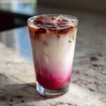 A tall glass showcasing the beautiful layers of an Iced Raspberry Vanilla Chai with bright pink syrup, white milk, and dark chai concentrate on ice.