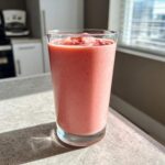 A tall glass filled with a thick, pink Iced Vanilla Strawberry Protein Shake sitting on a bright kitchen counter.