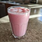 A tall glass filled with a frothy, pink Iced Vanilla Strawberry Protein Shake topped with ice and strawberry pieces.