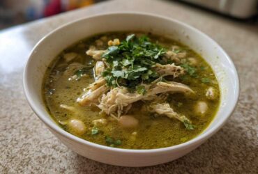 A close-up of a white bowl filled with vibrant green Instant Pot Chicken Pozole Verde, topped with shredded chicken and fresh cilantro.