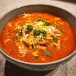 A close-up of a bowl filled with vibrant red Instant Pot Creamy Chicken Enchilada Soup, topped with shredded chicken, cheese, and cilantro.