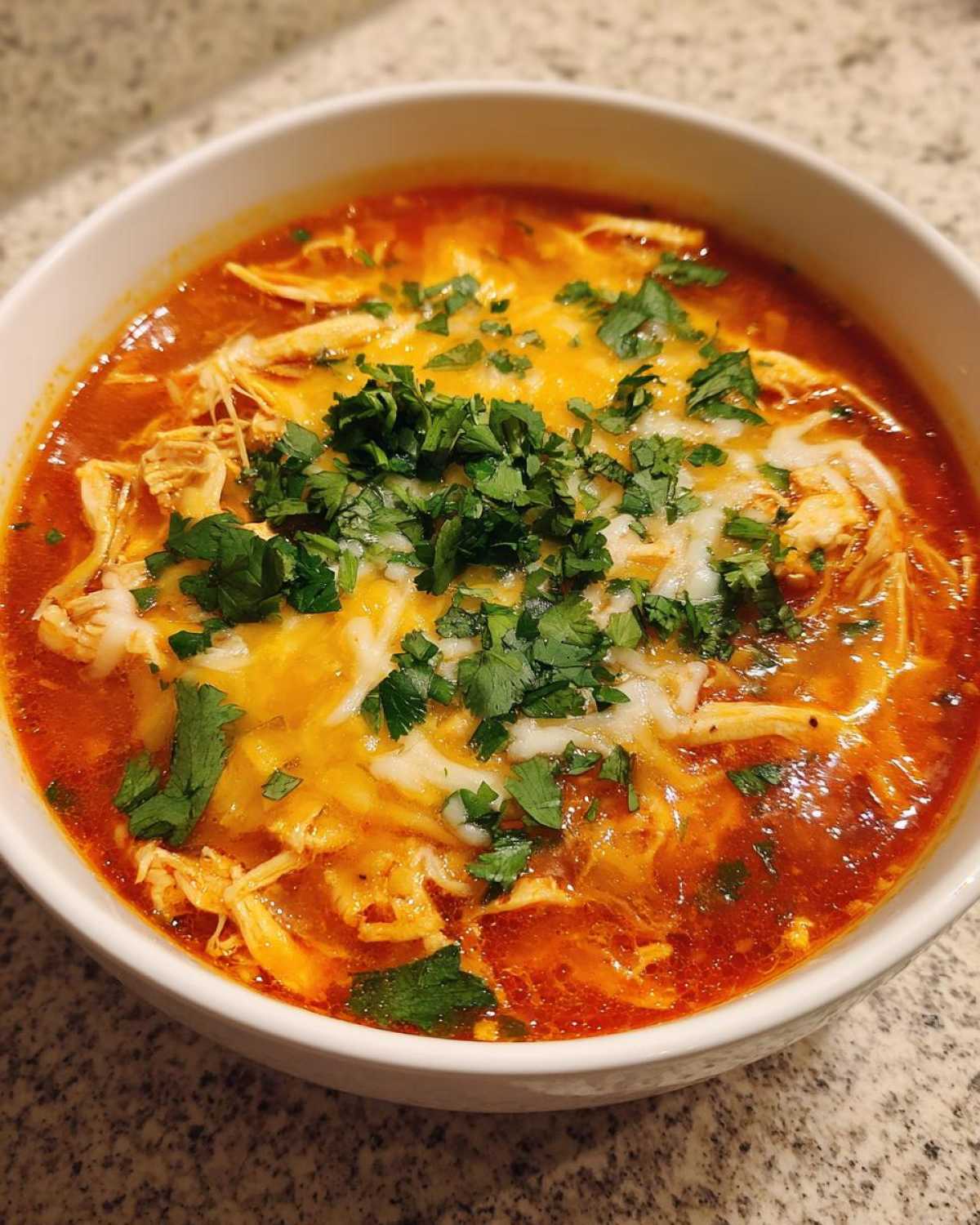 Close-up of a bowl of rich, red Instant Pot Creamy Chicken Enchilada Soup topped with melted cheese and fresh cilantro.