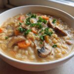 A close-up of a white bowl filled with creamy Instant Pot Creamy Mushroom Barley Soup, topped with sliced mushrooms and fresh parsley.