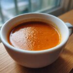 Close-up of a steaming white bowl filled with vibrant orange Instant Pot Creamy Red Pepper Tomato Soup, set on a wooden surface.