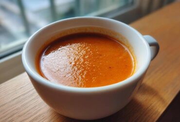 Close-up of a steaming white bowl filled with vibrant orange Instant Pot Creamy Red Pepper Tomato Soup, set on a wooden surface.