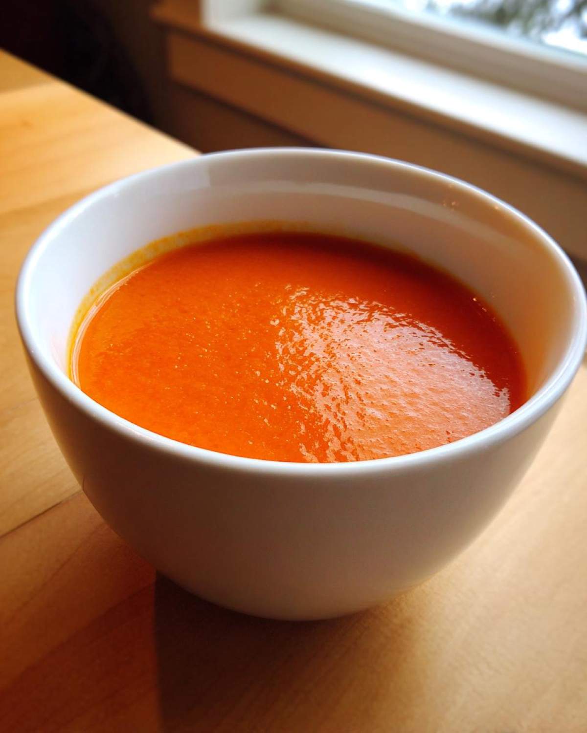 Close-up of a white bowl filled with vibrant orange Instant Pot Creamy Red Pepper Tomato Soup on a wooden surface.