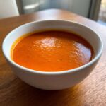Close-up of a white bowl filled with vibrant orange Instant Pot Creamy Red Pepper Tomato Soup on a wooden table.