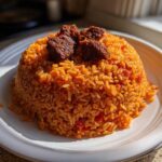 A mound of vibrant orange Jollof Rice served on a white plate, topped with pieces of dark, seasoned meat.