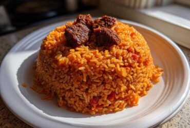 A mound of vibrant orange Jollof Rice served on a white plate, topped with pieces of dark, seasoned meat.