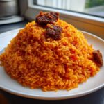 A mound of vibrant orange Jollof Rice topped with pieces of seasoned meat, showcasing the Irresistible Jollof Rice Flavor Adventure.