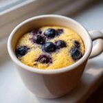 Close-up of a freshly baked Keto Blueberry Lemon Mug Cake with whole blueberries baked into the bright yellow cake.