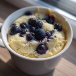 Close-up of a freshly baked Keto Blueberry Lemon Mug Cake topped with whole blueberries in a white mug.