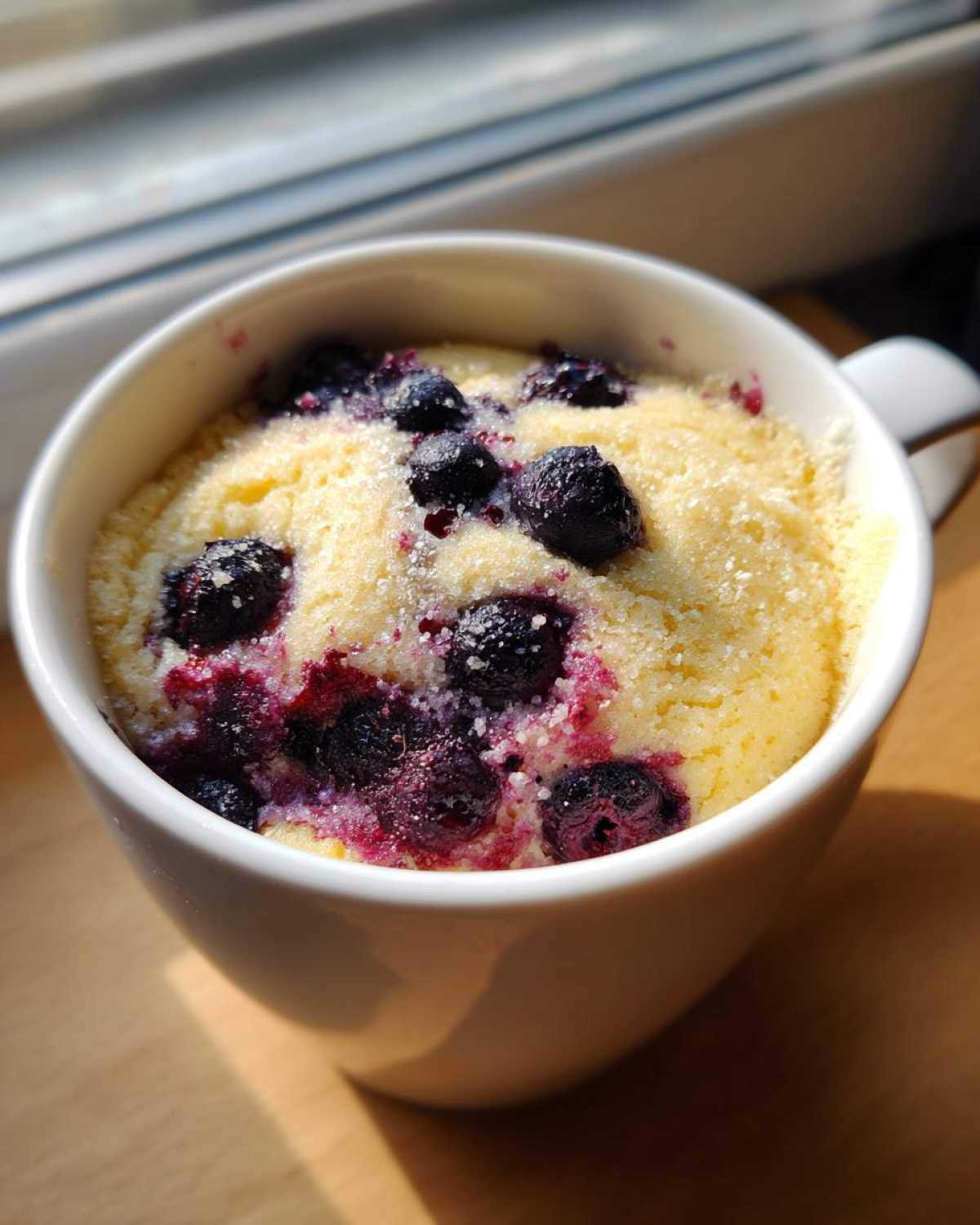 Close-up of a freshly baked Keto Blueberry Lemon Mug Cake topped with blueberries and sugar crystals in a white mug.