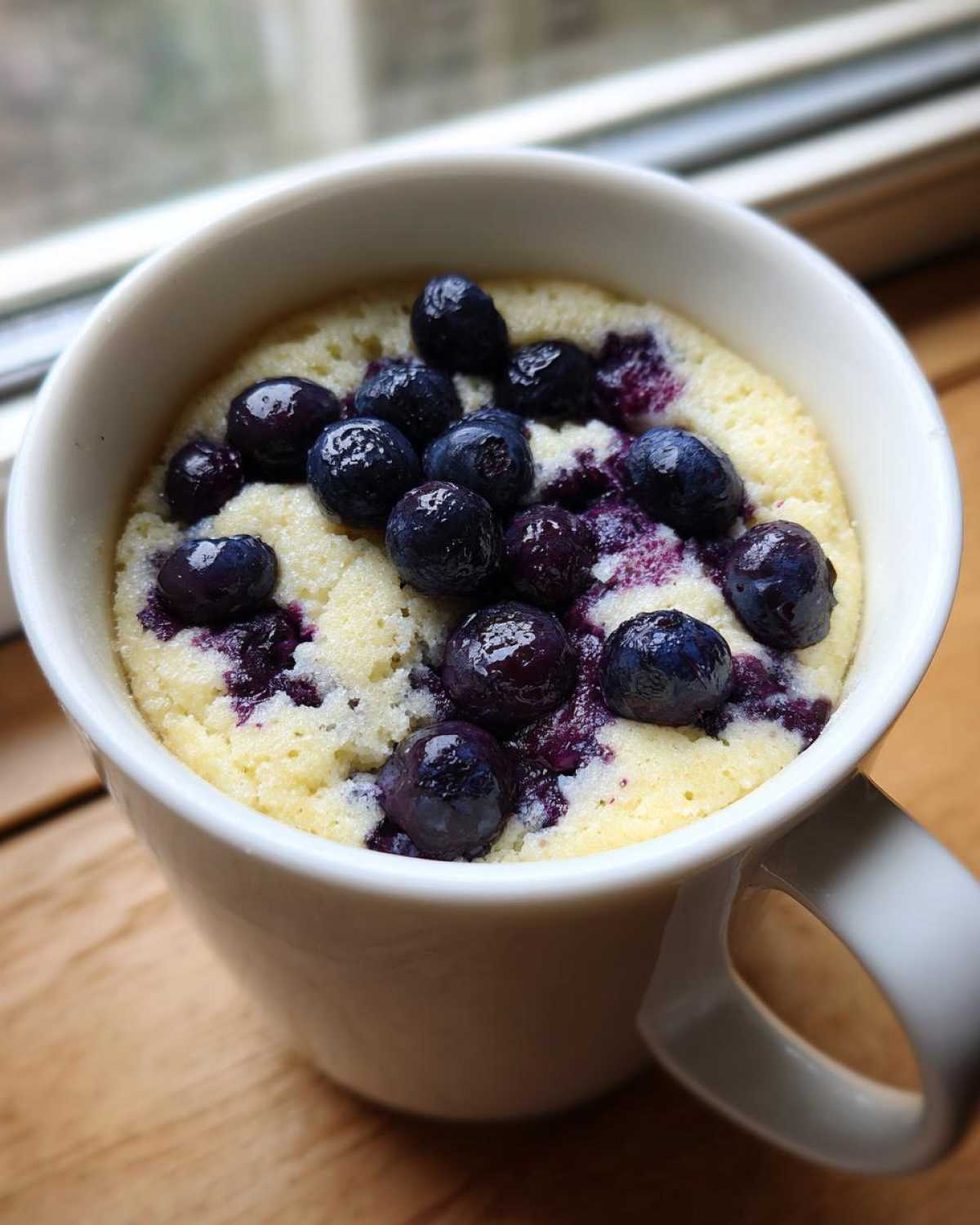 Close-up of a warm Keto Blueberry Lemon Mug Cake topped with fresh blueberries in a white mug.
