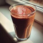 A tall glass filled with a thick, dark brown Keto Chocolate Strawberry Smoothie resting on a bright windowsill.
