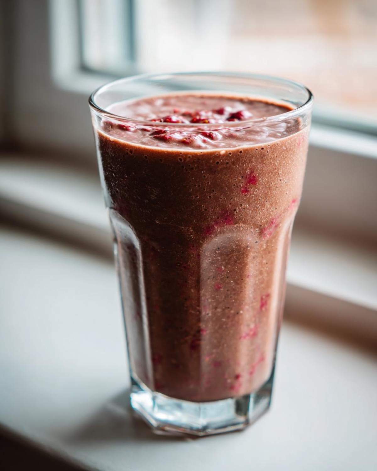 A tall glass filled with a thick, rich brown Keto Chocolate Strawberry Smoothie, showing flecks of red berry.