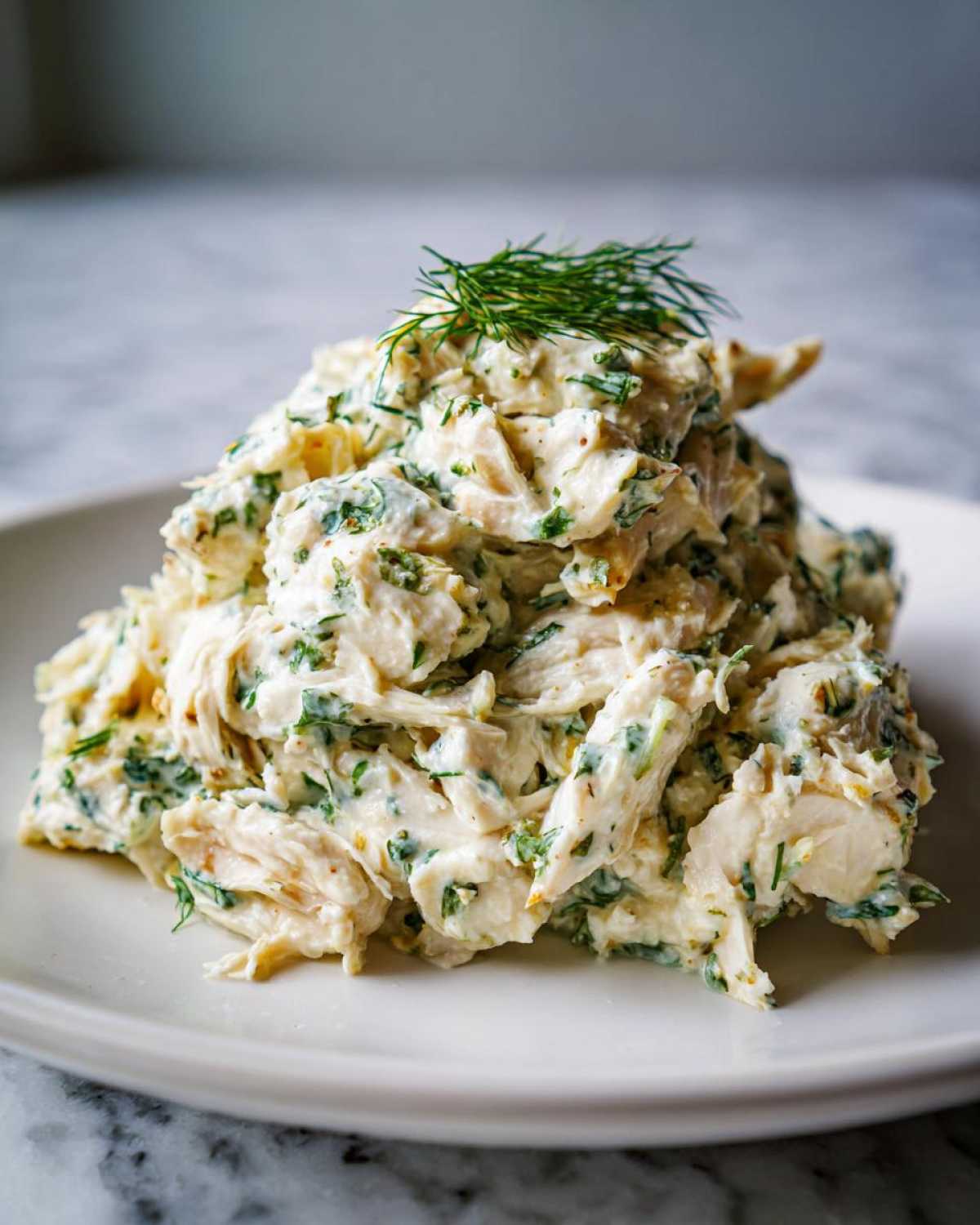 A mound of creamy Keto Lemon Herb Chicken Salad mixed with green herbs, garnished with fresh dill, served on a white plate.
