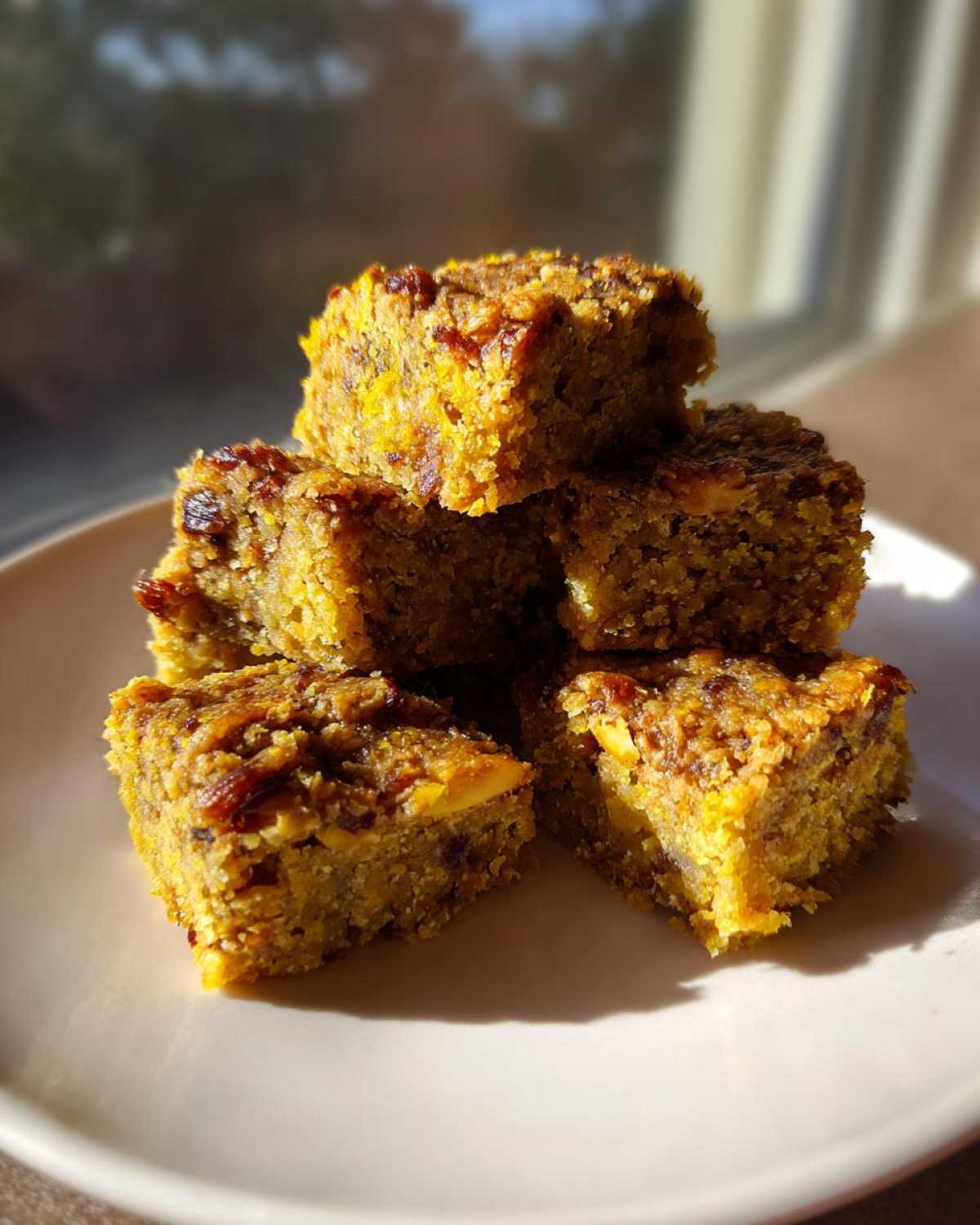 A stack of five square No-Bake Lemon Almond Date Bars resting on a white plate near a bright window.