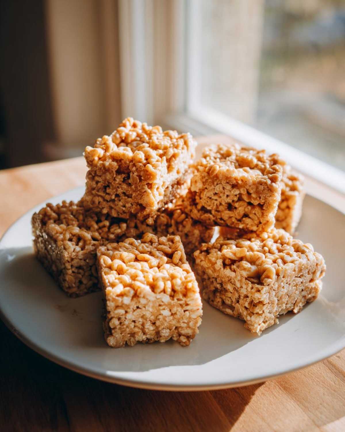 A stack of golden brown No-Bake Peanut Butter Rice Cereal Bars resting on a light plate near a window.