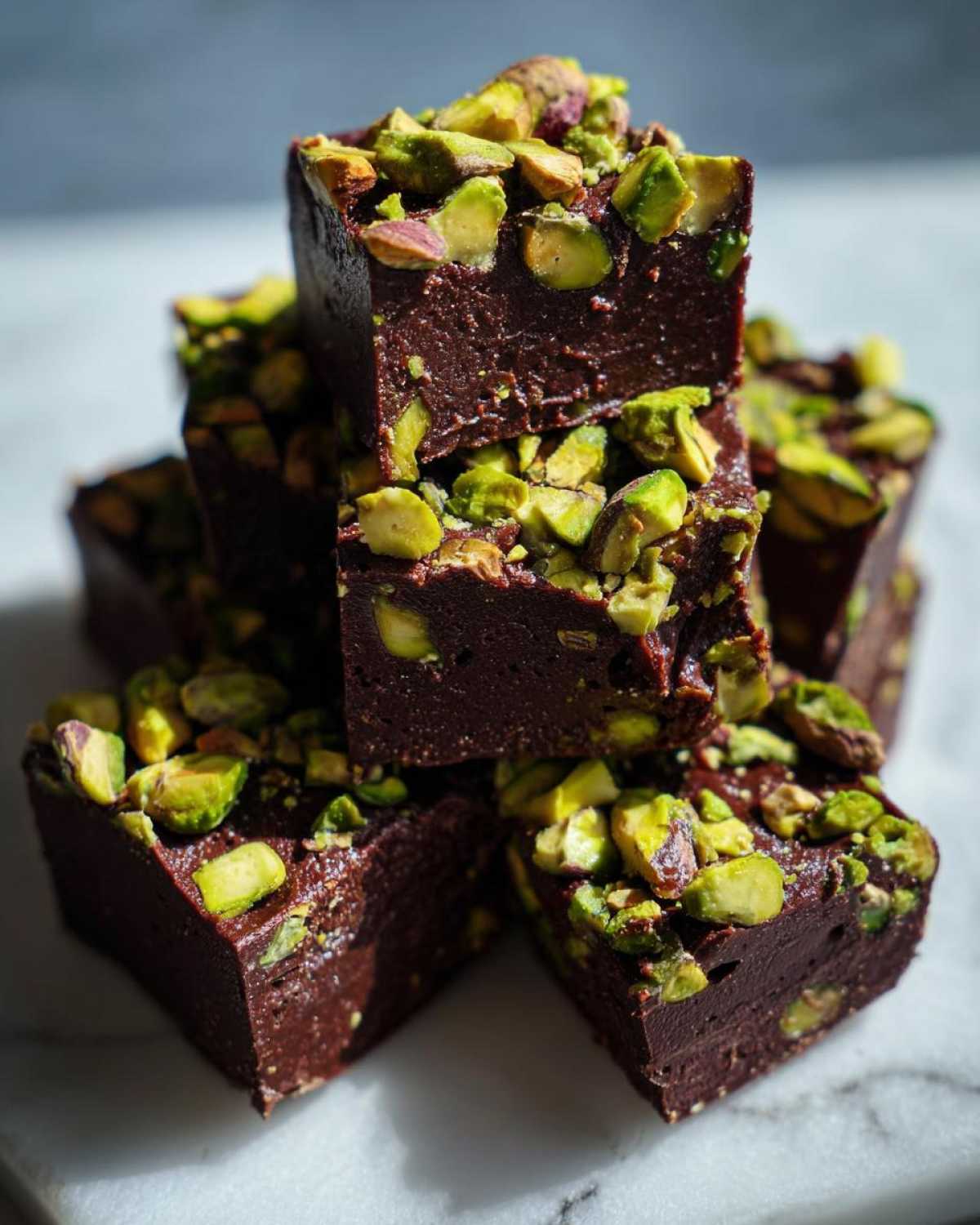 A stack of rich, dark chocolate No-Bake Pistachio Chocolate Fudge Squares topped with chopped bright green pistachios.