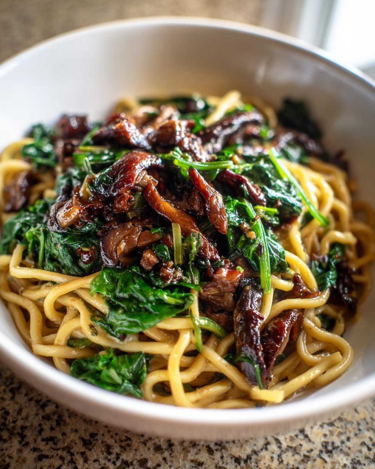 A close-up view of a white bowl filled with One-Pot Creamy Mushroom Spinach Ramen, showing noodles topped with dark mushrooms and bright green spinach.
