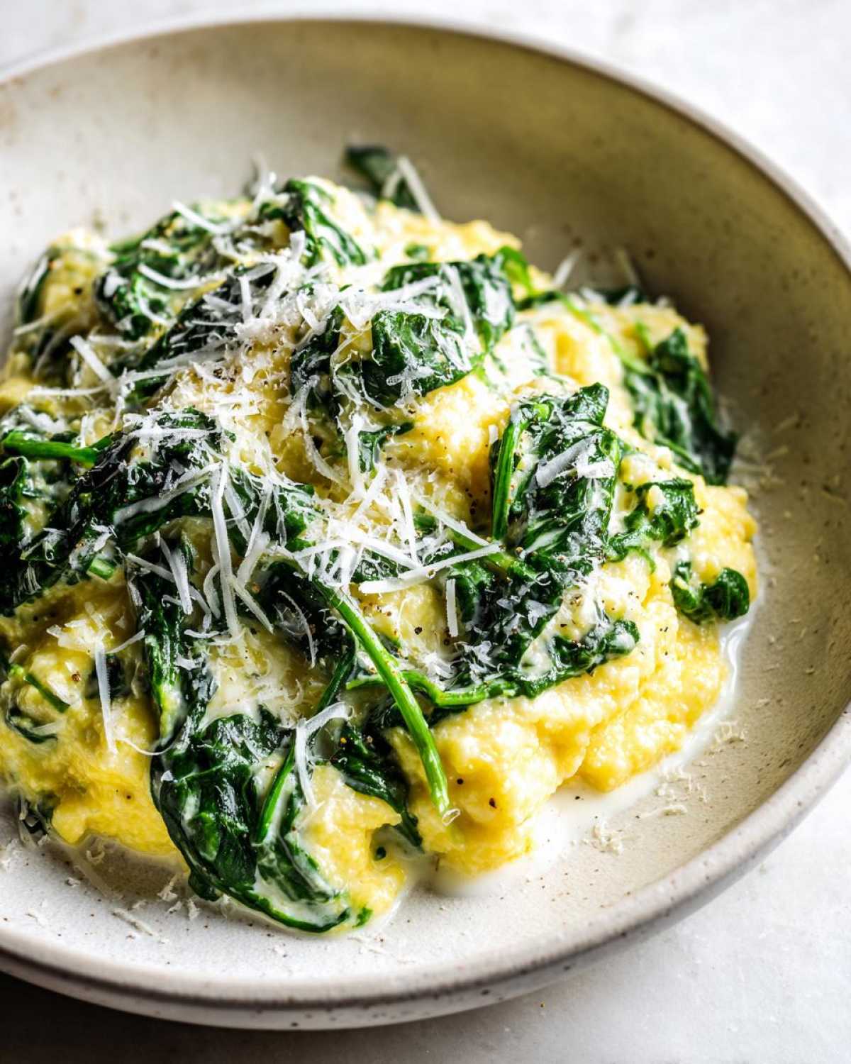 Close-up of One-Pot Creamy Spinach Parmesan Polenta topped with shredded Parmesan cheese in a beige bowl.