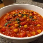 A close-up of a white bowl filled with rich, red One-Pot Creamy Tomato Basil Chickpea Stew, garnished with fresh basil.